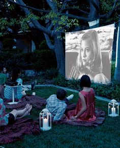 two people sitting on the grass watching a movie in front of a screen with candles