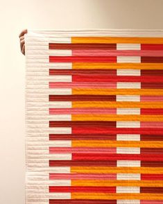 a woman holding up a quilt made from strips of red, orange and white fabric