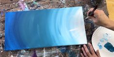 a person is painting on a canvas with blue and white colors, while another hand holds a paintbrush