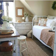 a white bed sitting under a window next to a wooden table with flowers on it