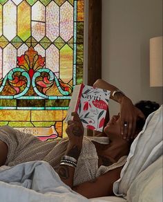 a woman laying in bed reading a book with a stained glass window behind her on the wall