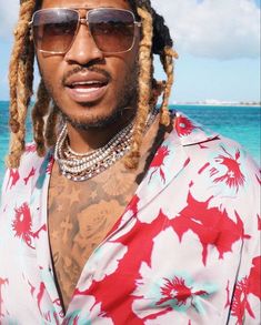 a man with dreadlocks standing in front of the ocean wearing sunglasses and a flower shirt