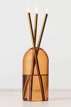 two candles in a glass vase with gold sticks sticking out of the top, on a white surface