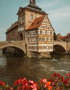 an old building sitting on the side of a river next to a bridge with flowers in front of it