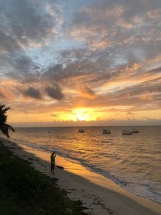 a person standing on the beach at sunset with boats in the water and people walking along the shore