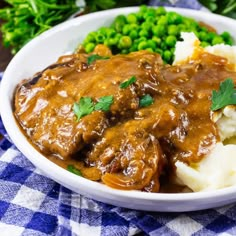 a white bowl filled with meat and gravy next to green peas on a blue checkered table cloth