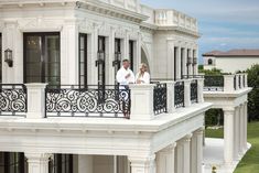 a man and woman standing on the balcony of a large white house with black iron railings