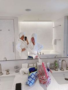 two women in white robes standing in front of a bathroom mirror taking a selfie