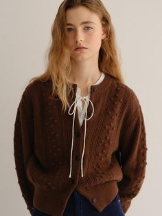 a woman wearing a brown cardigan sweater and blue jeans standing in front of a white wall