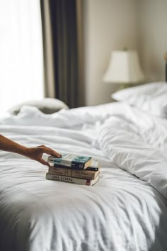 a person reaching for a stack of books on top of a bed with white sheets