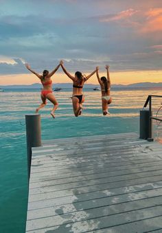 three women jumping off a dock into the ocean at sunset or dawn with their arms in the air