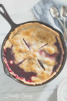 a blueberry pie in a cast iron skillet on a marble counter top with two spoons