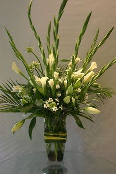 a glass vase with white flowers and greenery on a clear table in front of a wall