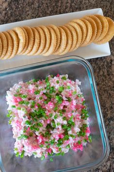 crackers are on the table next to a container with food in it and a bowl filled with rice sprinkled with green onions