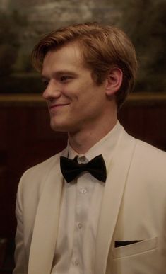 a man in a white suit and bow tie smiles at the camera as he looks off into the distance