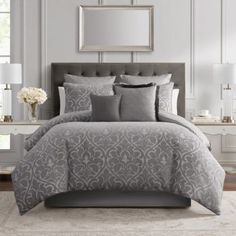 a bed with grey comforter and pillows in a room next to a mirror on the wall