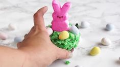 a hand is holding a small pink bunny in a nest filled with grass and eggs