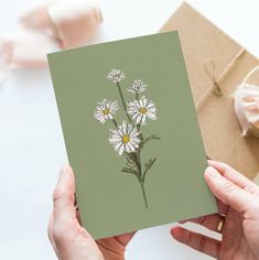 a hand holding a card with daisies on it and a gift box in the background