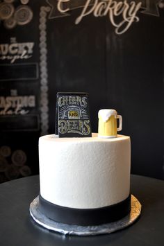 a beer themed cake with frosting and icing on the top is sitting on a table in front of a chalkboard wall