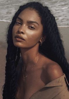 a woman with long black hair sitting on the beach looking off into the distance while wearing gold hoop earrings