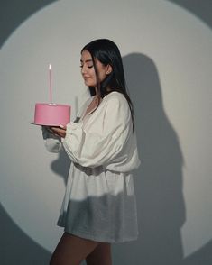 a woman holding a pink cake with a lit candle on it in front of a white wall