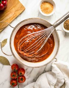 a bowl filled with sauce and whisk on top of a table next to tomatoes