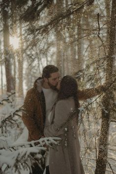 a man and woman standing in the snow near some trees with their arms around each other