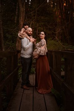 a man and woman are standing on a bridge in the woods with their baby girl