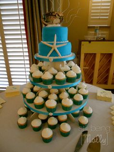 a blue and white tiered cake with cupcakes on the table next to it