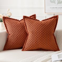 two orange pillows sitting on top of a white couch next to a vase with flowers