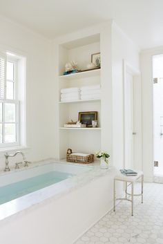 a white bathroom with a large bathtub next to a window and shelves on the wall