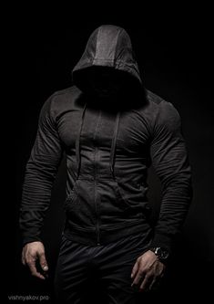 a man in black hoodie standing against a dark background with his hands on his hips