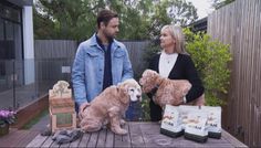 a man and woman standing next to two dogs on a wooden table with bags of dog food