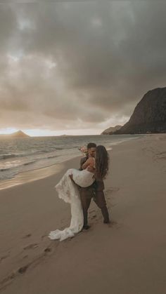 a bride and groom kissing on the beach in front of an overcast sky at sunset