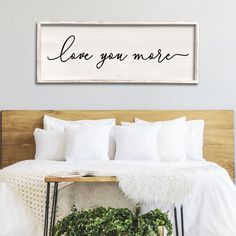 a bed with white sheets and pillows in front of a wooden sign that says love you more