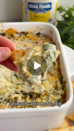 a hand holding a tortilla chip over a casserole dish filled with spinach and cheese