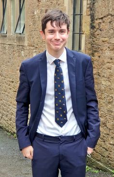 a young man wearing a suit and tie standing in front of an old building smiling for the camera