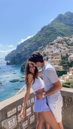 a man and woman standing next to each other in front of the ocean with mountains