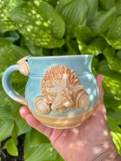 a hand holding a blue mug with a sea shell on the inside, and green leaves in the background