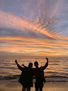 two people are standing on the beach with their arms in the air as the sun sets