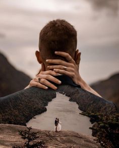 a man covering his face with both hands while standing on top of a mountain next to a river