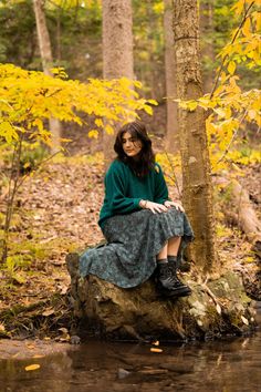 a woman sitting on top of a rock next to a tree in the woods with yellow leaves