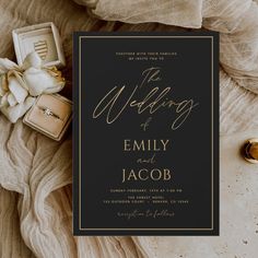 an elegant black and gold wedding card on top of a white cloth with two rings