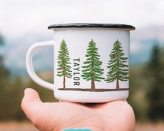 a hand holding a white coffee cup with trees on the front and bottom, in front of mountains