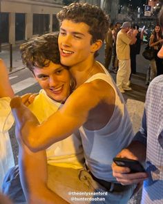 two young men hugging each other while standing on the street at night with people in the background