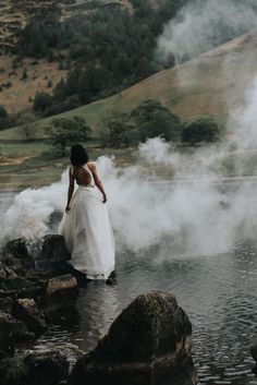 a woman in a white dress is standing on rocks by the water with steam coming from her back