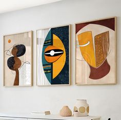 three abstract paintings hang on the wall above a dresser in a white room with two vases