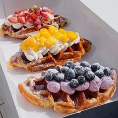 three waffles with different toppings on them sitting in a white box next to strawberries and blueberries