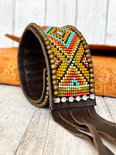 This bead loomed cuff bracelet is made from colorful glass seed beads in shades of mustard, olive, turquoise, terra cotta, orange, silver and black. The seed beads are hand loomed in a southwestern style design and sewn onto soft dark brown leather. Accented with brass beads and rhinestone chain. The beadwork is covered with a thin coat of resin to keep everything in place. The bracelet fastens around the wrist with a magnetic snap and features a long fringed piece of brown leather.  The cuff me Beaded Hat Bands, Bead Loom Designs, Beaded Leather Wraps, Beaded Memory Wire, Loom Bracelet Patterns, Beaded Hat, Beaded Cuff Bracelet, Bead Loom Bracelets, Brass Beads