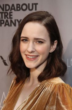 a woman with long brown hair smiling at the camera and wearing a gold dress in front of